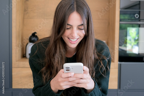 Smiling young woman in a cozy kitchen looking down at her smartphone, texting or browsing with a relaxed expression, long brown hair and casual sweater conveying modern home lifestyle and happiness