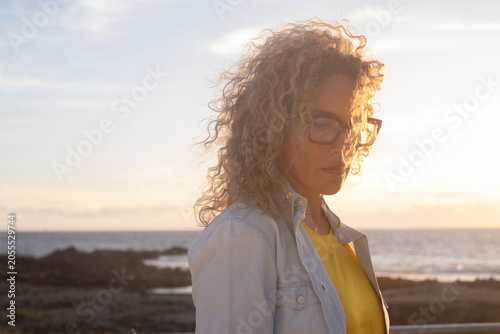 Contemplative woman with curly hair and glasses standing by the rocky beach at sunset, soft light and calm ocean in background
