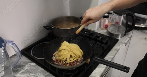 Chef adds freshly boiled spaghetti into frying pan filled with fried fat pork bacon. Rich savory aromas spread through warm kitchen. Slow motion