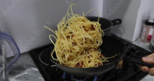 Chef mixes tender spaghetti with crispy bacon by shaking heavy black frying pan over stove. Aromatic steam rises through kitchen. Slow motion