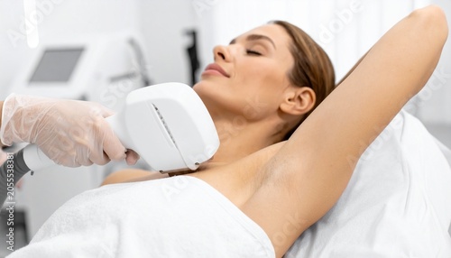 A woman undergoing laser hair removal treatment on her underarm in a brightly lit clinic. The technician's hand, wearing a glove, holds the laser device near her skin.