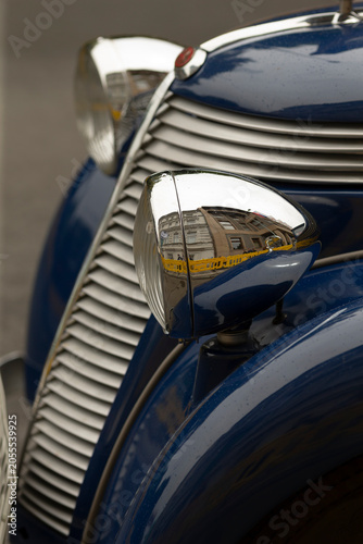 Detailed close-up of a polished chrome headlight and curved grille on the front fender of a blue classic automobile. The reflective chrome surface shows a