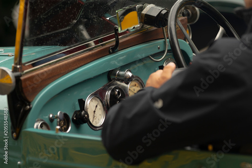 Photography Close-up view of a person steering a classic car featuring a turquoise painted dashboard, polished chrome instruments and round analog gauges