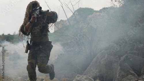 Photography A special forces soldier in a ghillie suit advances through a dense smoke screen on a rocky mountain, conducting a covert tactical military operation