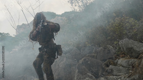 Photography A special forces soldier in a ghillie suit advances through a dense smoke screen on a rocky mountain, conducting a covert tactical military operation