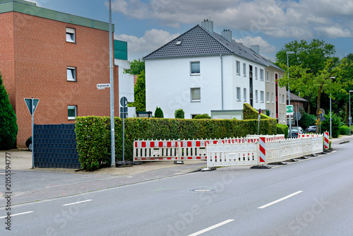 Obraz na plátně A street scene featuring modern residential buildings, green hedges, and temporary plastic safety barriers for roadworks or construction under a cloudy sky