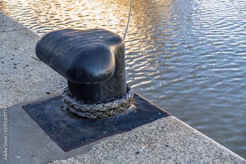 Schilderij op canvas Heavy-duty black metal mooring bollard on a concrete pier with a thick rope, ove