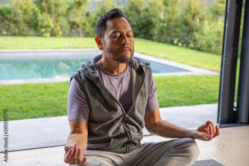 Mature Asian man meditating on yoga mat in activewear by sliding wall overlooking pool and lawn