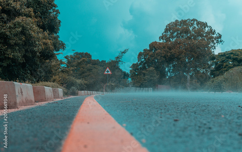 Obraz na plátně Misty wet paved road leading toward a triangular sign under a thick cloudy sky surrounded by large trees