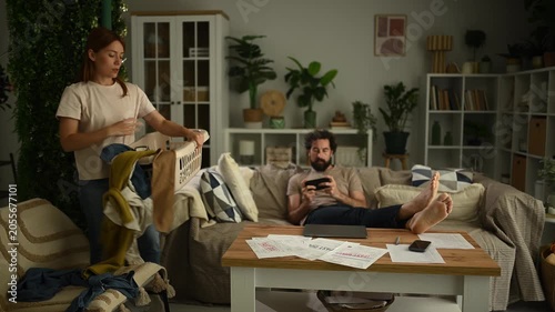 Frustrated woman doing housework and laundry while her irresponsible partner plays games on the sofa, ignoring financial problems