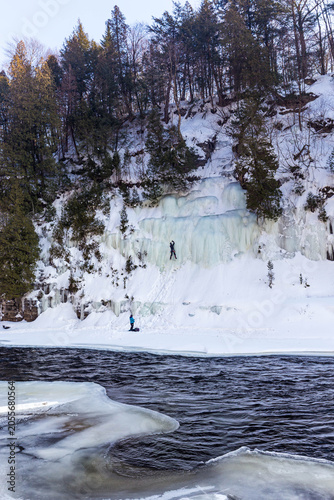 Ice climber ascending a frozen waterfall beside a river in a snowy forest, Quebec, Canada