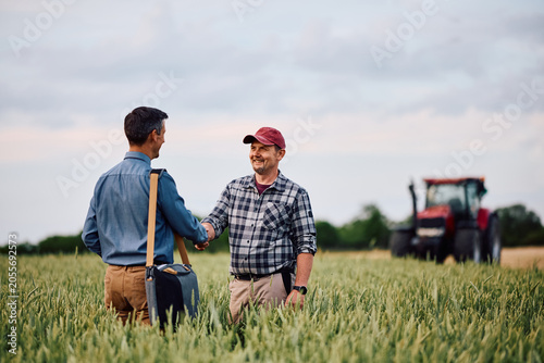 Happy agronomist shaking hands with his insurance consultant while standing in wheat field.