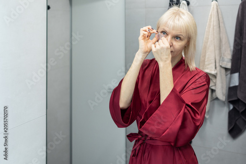 Patch Application Process. Blonde woman with short haircut in burgundy silk robe standing in bathroom before mirror while applying facial patches.