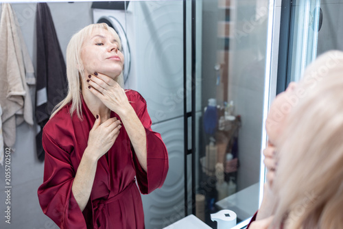 Skincare. Blonde woman with short hairstyle in burgundy silk robe doing facial massage in bathroom interior reflected in mirror.