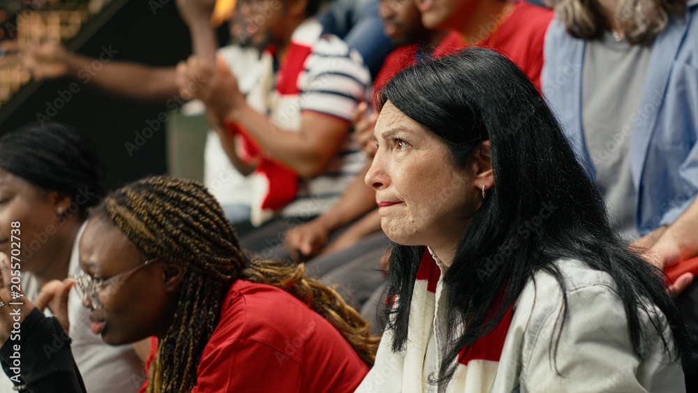 Fototapeta premium Red team football supporters reacting with anxious gestures in stadium stands during tense live soccer match, scarves and crowd emotion, fans with clenched fists during crucial moment in the match.