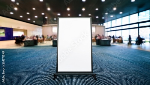 blank vertical easel poster in modern indoor event space with blurred exhibition backdrop ideal for conference signage business presentation or commercial promotional mockup