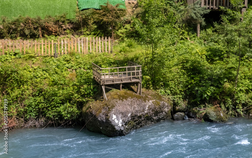 Wooden lookout structure built on a large mossy boulder overlooking a turquoise mountain river. The scene is surrounded by dense green forest. Evoking a serene and rustic atmosphere.