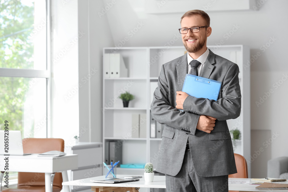 Naklejka premium Smiling young businessman in gray suit holding blue clipboard in modern office environment, he wears glasses and has a beard, exuding confidence and professionalism