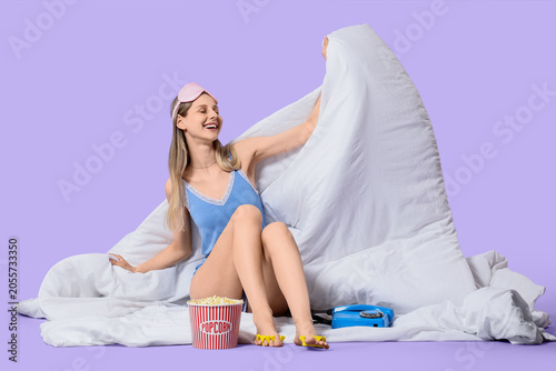 Young woman in pajamas with popcorn and telephone sitting on white blanket ag...
