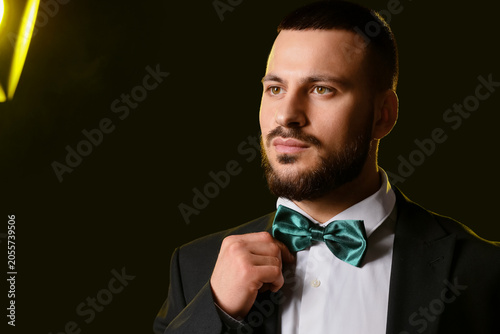 Young man in stylish suit adjusting bow tie on dark background, closeup