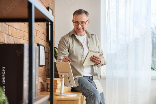 Smiling mature man using digital tablet and notebook for remote work or onlin...