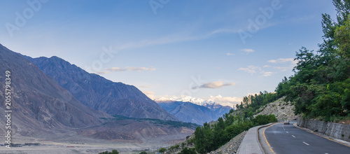 Foto Panoramic view of a winding road covered by lush greenery contrasting with harsh Karakoram mountains and distant snow peaks