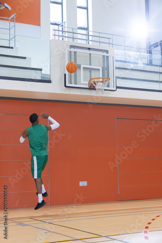 Basketball is arcing toward glass backboard and rim in gymnasium with orange wall