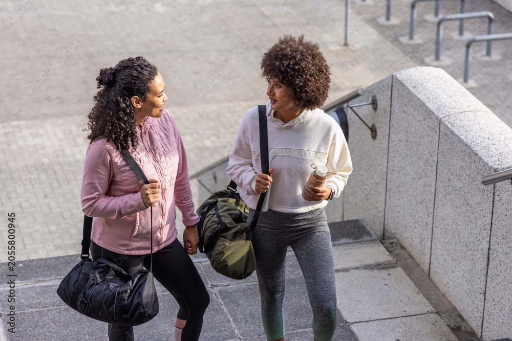 Fototapeta premium Two female friends in workout clothes walking up stairway at plaza carrying duffel bags and bottle