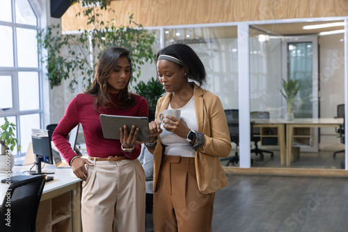 Indian and African American businesswomen standing in open office, viewing tablet and holding mug