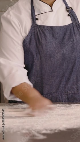 Vertical video: Reaching, chef spreading flour on metal counter for dough prep, wearing denim apron