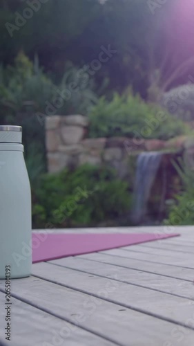 Vertical video: Camera panning, bringing water bottle onto wooden deck near pink yoga mat for yoga
