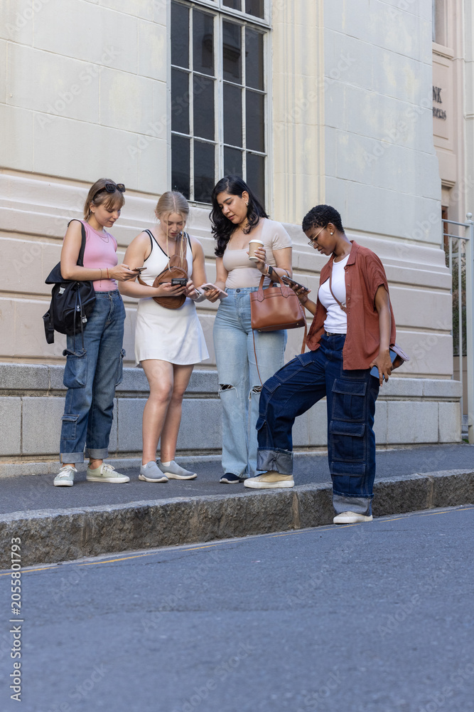 Fototapeta premium Diverse female friends standing on stone curb, checking smartphones and holding takeaway coffee cup