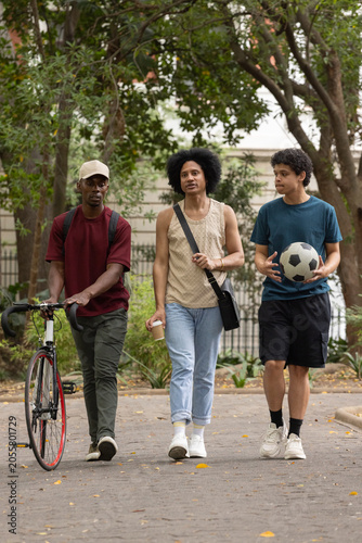 African American males walking in park pushing bike with coffee, teenage male holding soccer ball