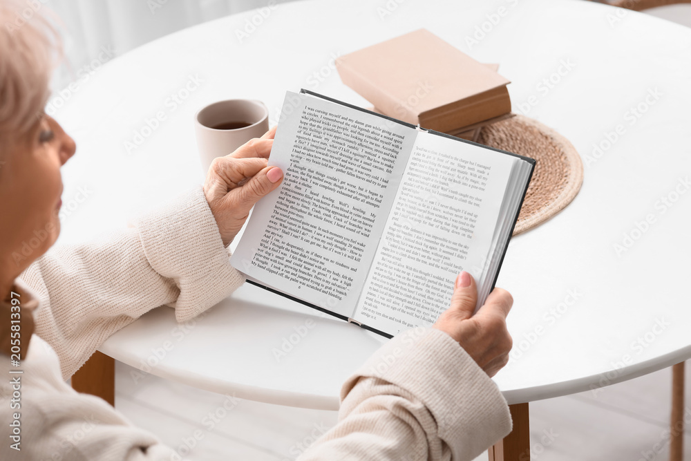 Fototapeta premium Beautiful senior woman reading book at table in kitchen