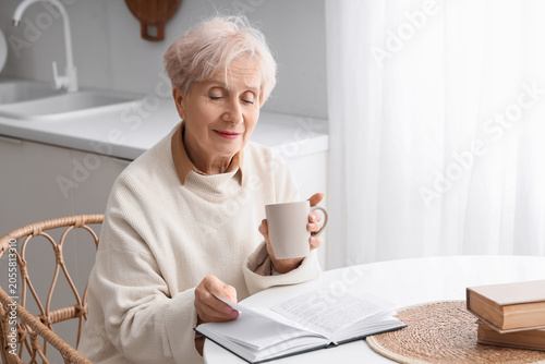 Schilderij op canvas Beautiful senior woman with tea cup reading book at table in kitchen
