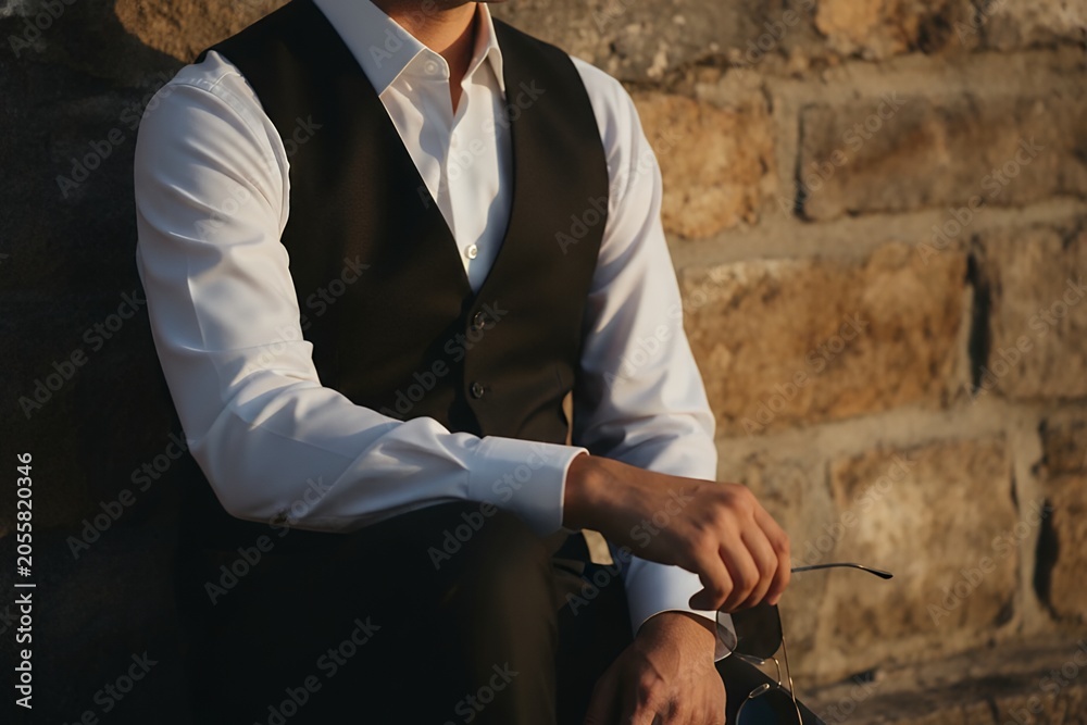 Fototapeta premium Man in vest and white shirt sits against stone wall with glasses