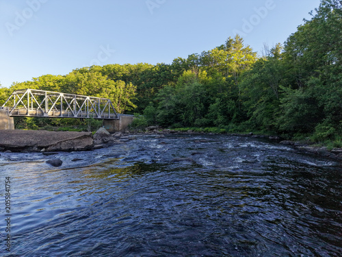 Εκτύπωση καμβά A peaceful riverscape featuring a steel truss bridge spanning flowing water with rocky banks and dense green forest under a clear summer sky