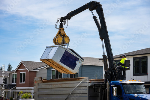 Construction dumpster being emptied by knuckle boom arm on specialized grapple truck, operator in control seat, new residential housing development
