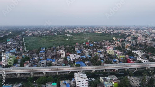 An impressive top-down drone perspective tracking the sleek line of a metro railway line cutting across a bustling Kolkata neighborhood, emphasizing eco-friendly public transportation infrastructure