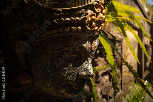 Ancient carved stone deity sculpture with a serene expression, representing Hindu spirituality and culture in an old Bali temple garden, illuminated by sunlight and shadows