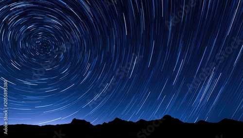 Swirling star trails above a dark, silhouetted mountain range under a deep blue night sky