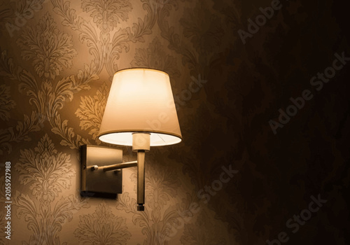 Modern brass wall sconce with cream fabric lampshade mounted on textured damask patterned wallpaper, providing warm ambient lighting in a dark elegant interior room setting.