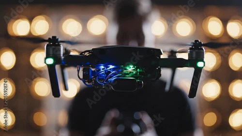 Professional quadcopter drone hovering in a dimly lit studio with warm bokeh background lights and a pilot in the background