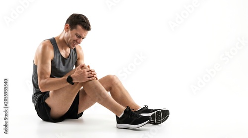 Young athletic man sitting on a white background and holding his injured knee in pain after exercise.