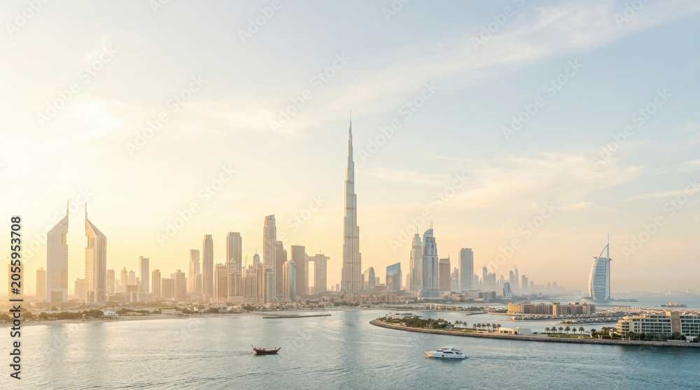 Fototapeta premium Dubai skyline skyscraper waterfront sunrise cityscape marina, calm harbor with modern tower, boat, island, and soft haze over coastal urban architecture in warm light