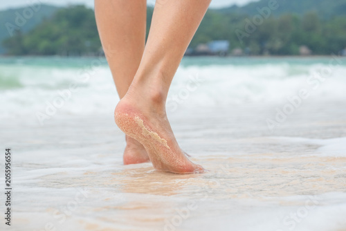 Close-up of a woman bare legs and feet walking away on a sandy beach. Sand adheres to the wet soles and heels. Waves, the ocean, and a green, tree-covered island are visible in the background