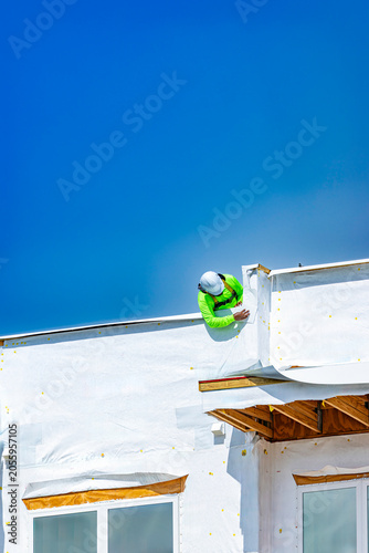A vertical view of a construction worker wearing a white hard hat, a bright neon green safety shirt, and a safety harness, leaning over the roof edge to install white weather-resistant housewrap barri
