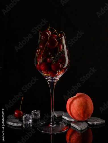 Still life. A large glass filled with cherries, a peach, and a cherry berry on stones. Black background.