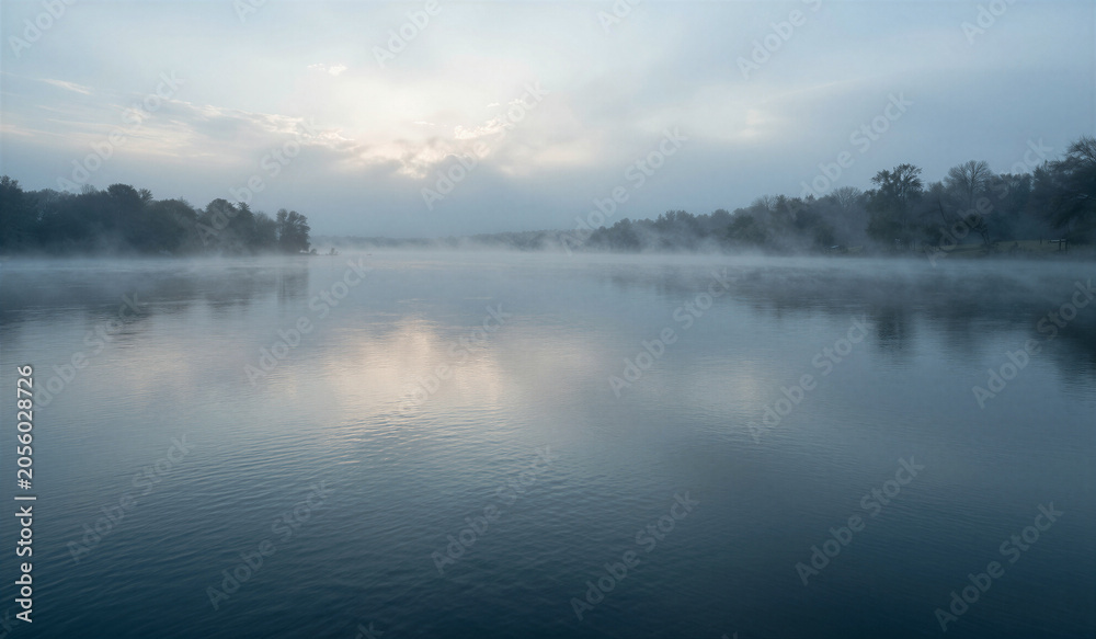 Fototapeta premium Mist clings to still water under soft dawn light. Trees stand hushed at lake's edge, veiled in fog. Sky bleeds pale blue and gray, fading toward horizon. Silent peace holds the scene - no sound