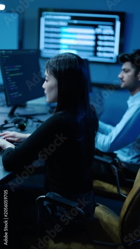 Professional IT experts work in a modern data center, managing software development, cybersecurity, and server network monitoring on multiple screens with complex code in a dark office environment.